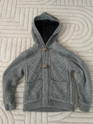 Grå stickad cardigan  - Riktigt fet grå stickad cardigan med träknappar och två framfickor. Tröjan har ett tydligt mönster i stickningen och ribbade muddar vid ärmslut och nederkant. Perfekt för lager på lager och chill vardagsstil. Modellen är 175 och väger 60kg 