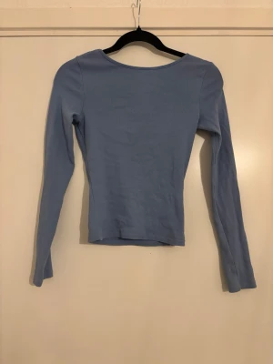 Ljusblå långärmad topp med djup rygg - Ljusblå långärmad topp med tight passform och djup, rundad rygg. Tillverkad i ett mjukt, ribbat material som sitter skönt mot kroppen. Perfekt för dig som gillar stilrena och enkla plagg med en twist.