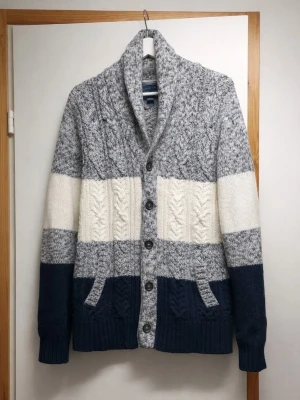 Stickad kofta med blockfärger - Mysig stickad kofta med breda blockränder i grått, vitt och marinblått. Koftan har grovstickat mönster, stora knappar framtill, två fickor och en hög krage. Perfekt för kalla dagar och ger en avslappnad men snygg look.