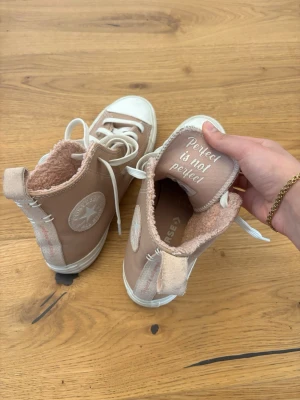 Beige Converse sneakers med foder - Säljer ett par beige Converse high top sneakers med vitt foder i teddy, vita snören och tåhätta. Klassisk Converse-logga på sidan och texten 'Perfect is not perfect' på plösen. Skorna har en chunky vit sula och är perfekta för en cool streetstil. Passar även mig med 37,5