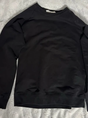 Svart Stone Island tröja - En enkel och stilren svart sweatshirt med rund hals och långa ärmar. Tröjan har ribbade muddar vid ärmslut och nederkant, perfekt för en avslappnad look. Materialet är mjukt och bekvämt