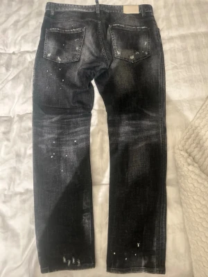 Svarta jeans från Dsquared2, stl 50 - Snygga svarta jeans från Dsquared2 med coola slitningar och vita färgstänk. Raka ben och klassisk femficksmodell. Jeansen har en unik tvättad look och är tillverkade i bomullsblandning. Köpta för 6300kr