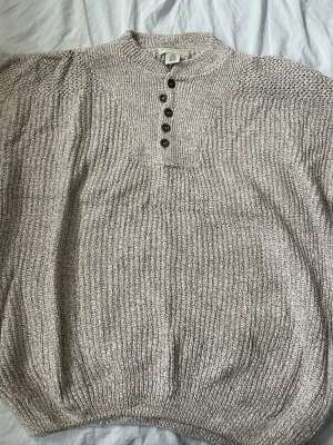 Beige stickad tröja från Eddie Bauer - Säljer en beige stickad tröja från Eddie Bauer med grovstickad struktur och rund hals. Tröjan har ett metallmärke med logotypen på nederkanten och är tillverkad i ett mjukt garn som ger en avslappnad look. Perfekt för lager-på-lager under kyliga dagar.