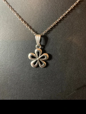 Silverhalsband med blomma - Snyggt halsband i silverfärg med en hängande blomma som har fem rundade kronblad. Kedjan är tunn och smidig, och blomman har en modern, stilren design. Perfekt för dig som gillar enkla men unika smycken. Märke och material är ej angivet.