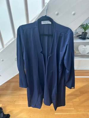 Mörkblå tunn kapla  - Stilren mörkblå kappa med öppen front och knytskärp i midjan. Kappan har trekvartsärmar och en rak, avslappnad passform som ger en modern look. Perfekt för lager på lager och enkel att matcha till olika outfits.