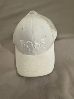 Vit keps från Hugo Boss - Snygg vit keps från Hugo Boss med broderad logga framtill. Klassisk böjd skärm och justerbar rem bak. Tillverkad i bomullstyg som ger en clean och stilren look. Perfekt accessoar för en casual streetwear-stil. Har en liten fläck men går säkert bort om man tvättar den.