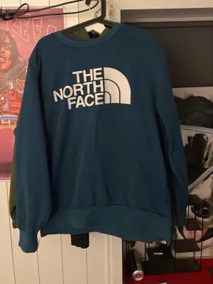 Blå sweatshirt från The North Face - Snygg blå sweatshirt från The North Face med stort vitt logotryck på framsidan och mindre logga på ryggen. Tröjan har rund halsringning och ribbade muddar vid ärmslut och nederkant. Perfekt för en avslappnad och cool stil.
