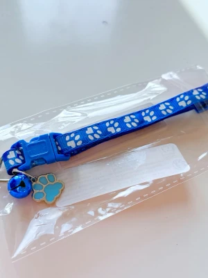 Blått katt-halsband med tassmönster - Snyggt blått katt-halsband i nylon med vita tassavtryck, ett blått pinglande bjällra och en söt tassformad berlock. Halsbandet har ett justerbart spänne i plast och passar perfekt till din katt eller lilla hund.