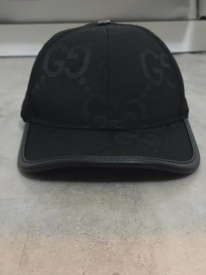 Svart keps från Gci med GG-mönster - Snygg svart keps från Gci med diskret GG-mönster över hela kepsen. Kepsen är tillverkad i bomull och polyester med en klassisk böjd skärm och stilren design. Perfekt accessoar för att lyfta din streetwear-look.