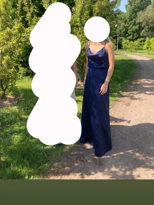 Marinblå långklänning med smala axelband - Elegant marinblå balkläning med smala axelband och v-ringad urringning. Klänningen har en stilren, figurnära passform och ett mjukt, glansigt tyg som ger en lyxig känsla. 