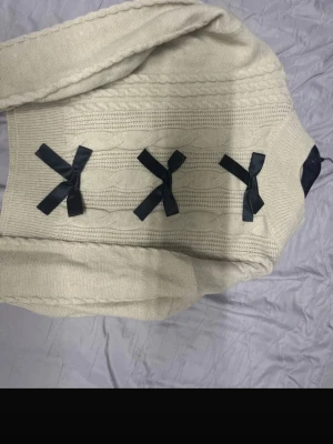 Beige stickad tröja med svarta rosetter - Supersöt beige stickad tröja med kabelstickat mönster och tre svarta rosetter på ryggen. Tröjan har långa ärmar och ribbade muddar. Perfekt för dig som gillar gulliga detaljer och en mysig stil.