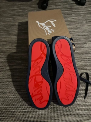 Svarta sneakers från Christian Louboutin - Snygga svarta sneakers från Christian Louboutin med klassisk röd sula och signaturdetaljer undertill. Skorna har rund tå, snörning och är tillverkade i mocka med mjuk beige insida. Perfekt för dig som vill sticka ut med exklusiv stil. Box ingår ej