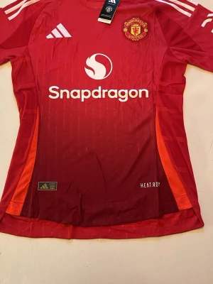 Manchester United matchtröja röd Adidas - Snygg röd Manchester United matchtröja från Adidas med vita detaljer och klubbmärke på bröstet. Tröjan har Snapdragons logga framtill, vita ränder på axlarna och är tillverkad i ett lätt, tekniskt material med HEAT.RDY-teknologi. Perfekt för fotbollsträning eller match.