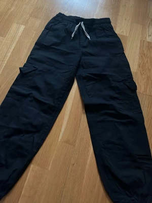 Svarta cargobyxor med dragsko - Säljer ett par svarta cargobyxor med elastisk midja och vit dragsko. Byxorna har stora sidofickor med lock och dragsko vid bensluten för en avslappnad look. Perfekta för en streetwear-stil och tillverkade i ett mjukt bomullsliknande material.
