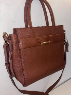 Brun handväska i läder från Hera - Snygg brun handväska från Hera i äkta läder med struktur. Väskan har både handtag och justerbar axelrem, samt guldfärgade metalldetaljer. Klassisk rektangulär form och en stilren design med diskret logga framtill.