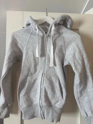 Russemerch zip-hoodie  - En klassisk ljusgrå hoodie med dragkedja och huva. Tröjan har stora fickor framtill, breda muddar vid ärmslut och nederkant samt vita snören i huvan. Perfekt för en avslappnad och sportig stil.