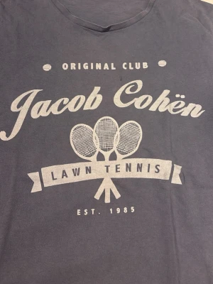 Blå t-shirt Jacob Cohën  - Snygg blå t-shirt från Jacob Cohën med tryck 'Lawn Tennis' och tre tennisrack på bröstet. T-shirten har rund hals och är tillverkad i mjuk bomull. Storlek M, jätte bra skick, ny pris 1800
