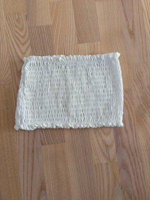 Vit bandeau tubtopp - Säljer en vit bandeau tubtopp med smockad resår runtom. Toppen är ärmlös och har en enkel, clean look som passar perfekt till sommaren. Materialet känns mjukt och stretchigt för en bekväm passform. Passar även M