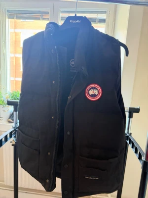 Svart dunväst från Canada Goose - Svart dunväst från Canada Goose med klassisk logotyp på bröstet och två stora fickor framtill. Västen har hög krage, tryckknappar och dragkedja. Perfekt för lager-på-lager och håller dig varm under kyliga dagar, Västen har ett litet hål inuti, skriv för mer bilder