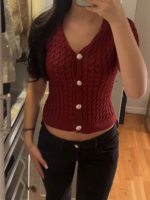 Vinröd kabelstickad kortärmad kofta - Snygg vinröd kabelstickad kofta med korta ärmar och djup v-ringning. Koftan har stora vita knappar framtill och är croppad i modellen. Perfekt att styla med jeans eller kjol för en trendig look.