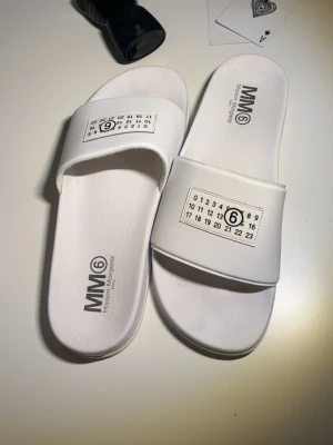 Vita slides från MM6 Maison Margiela - Snygga vita sandaler från MM6 Maison Margiela med ikonisk logotyp och siffror på remmen. Ändast haft dom på mig 7 gånger. Tillverkade i syntetmaterial med bekväm sula och stilren design. 