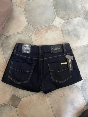 Mörkblå lågmidjade denimshorts från Goodies - Snygga mörkblå denimshorts från Goodies Jeans med kontrastsömmar och två bakfickor. Storlek 36, vilket motsvarar S. Klassisk femficksmodell med låg midja och tydlig logga bak. Perfekta för en trendig och avslappnad stil.