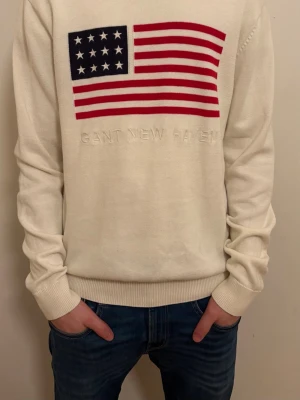 Vit gant tröja - Säljer en stilren vit stickad tröja från GANT med en amerikansk flagga och texten 'GANT NEW HAVEN' på framsidan. Tröjan har långa ärmar och en rund halsringning. Perfekt för en avslappnad och trendig look. Lappen sitter löst men går säkert att fixa, storlek M