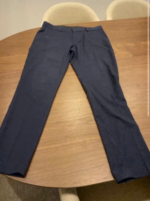 Mörkblå kostymbyxor Isaac Dewhirst - Snygga mörkblå kostymbyxor från Isaac Dewhirst i klassisk rak modell. Byxorna har bälteshällor och är tillverkade i ett slätt tyg som ger en stilren look. Perfekta för dig som vill ha en clean och modern stil.