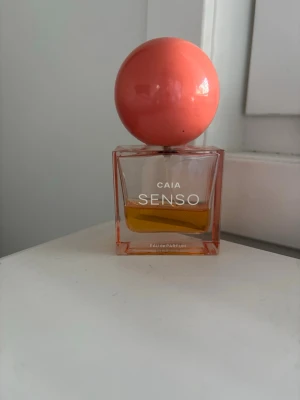 CAIA Senso Eau de Parfum - Parfymflaska från CAIA med namnet Senso. Flaskan är fyrkantig i genomskinligt glas med en stor, rund, korallfärgad kork. Innehållet är gyllene gult och designen känns modern och trendig. Perfekt för dig som gillar snygga detaljer på parfymhyllan. Jättefin sommardoft 