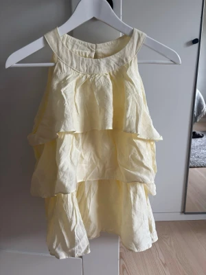 Gul volangtop med halterneck - Säljer en ljusgul ärmlös topp med halterneck och flera volanglager. Toppen är aldrig använd, därav ny skick
