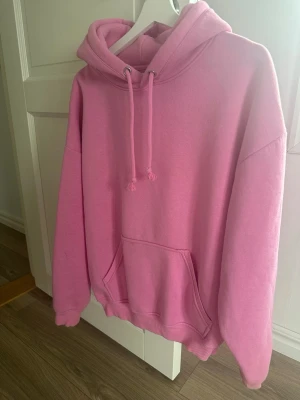 Rosa hoodie med huva och magficka - Säljer en rosa hoodie med klassisk huva och dragsnören. Hoodien har en stor magficka framtill och ribbade muddar vid ärmslut och nederkant. Perfekt för en chill och avslappnad stil. Materialet känns mjukt och skönt, troligen bomullsblandning.