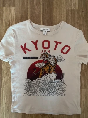 Vit t-shirt med tigertryck från Topshop - Snygg beige t-shirt från Topshop med stort tryck av en tiger, röd sol och texten KYOTO på framsidan. T-shirten har korta ärmar och rund hals. Perfekt för dig som gillar streetstyle och coola prints.