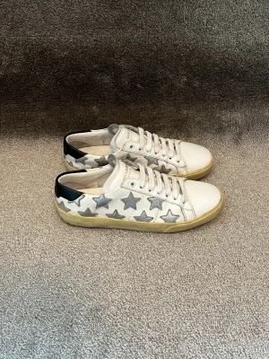 Saint Laurent skor - Hej! Säljer dessa väldigt fina Saint Laurent skor i ett väldigt gott skick! Skorna har silverstjärnor runt om och svart läder detalj bak vid hälen på skorna. Dessa kostar 7000kr nya! Box och extrasnören medföljer i köpet! Skorna är handgjorda i Spanien och är tillverkade i kalvskinn! Storleken är 36,5. Dessa är självklart äkta vilket jag står för! Det är olagligt att sälja märkeskopior. Produkt kod står stämplat i skorna och går att checkas i butik. Mvh DS