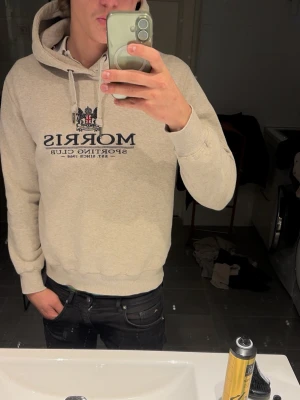 Beige hoodie från Morris Sporting Club - Snygg beige hoodie från Morris Sporting Club med broderad logga och text på bröstet. Klassisk huva utan zip och midja. Perfekt för en avslappnad och stilren look. Använd sällan och är i bra skick. Pris kan diskuteras!!!