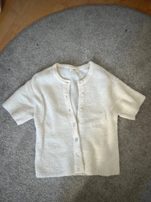 Vit kortärmad kofta från Arket - Säljer en krämvit, ribbstickad kofta från Gina med korta ärmar och rund halsringning. Koftan har knappar framtill och är gjord i ett mjukt, ull-liknande material. Perfekt att styla över ett linne eller t-shirt.