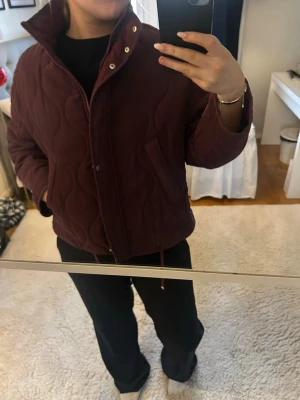 Mörkröd quiltad jacka med hög krage - Quiltad mörkröd jacka med hög krage och knappar framtill. Jackan har ett loose fit och är långärmad, perfekt för höst och vinter. Materialet känns som polyester och jackan har snygga sömmar som ger ett coolt mönster. Den är köpt på zalando. 
