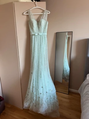 Ljusblå långklänning med paljetter från Dressroom Linne - Magisk ljusblå långklänning med v-ringning och breda axelband. Klänningen är täckt av glittriga paljetter och har ett tunt band i midjan. Kjolen är lätt utsvängd och dekorerad med små blommor i tyget. Perfekt för bal eller speciella tillfällen. 