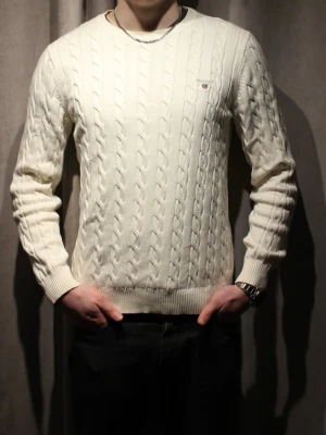 Vit kabelstickad tröja från Gant - Kabelstickad vit tröja från Gant med rund halsringning och diskret logga broderad på bröstet. Tröjan har ribbade muddar vid ärmslut och nederkant, vilket ger en klassisk och stilren look. Tröjan har inga defekter och ser splitterny ut