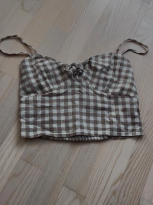 Beige och vit rutig linnetopp - Säljer en croppad linnetopp med beige och vitt rutigt mönster. Toppen har tunna axelband, knyt framtill och smockad rygg för extra komfort. Perfekt för varma dagar och enkel att styla med jeans eller kjol.