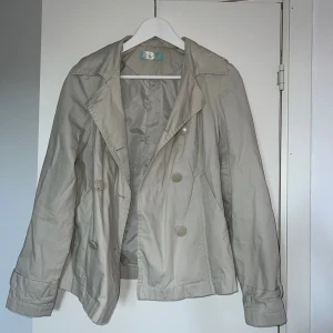 Beige trenchcoat från Thl - Stilren beige trenchcoat från Thl med dubbelknäppning och klassisk krage. Jackan har långa ärmar och en lätt glansig finish. Perfekt för dig som vill ha en tidlös och enkel look. Passar till många olika outfits och är ett måste i garderoben.