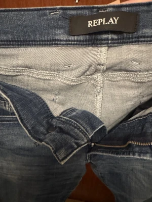 Blå jeans från Replay - Säljer ett par blå jeans från Replay med klassisk femficksmodell och dragkedjegylf. Jeansen har en snygg tvätt med slitningar och kontrastsömmar. Märkesdetaljer syns på linningen och fickan. Perfekta för en trendig och avslappnad stil.