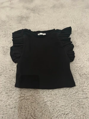 Svart topp med volangärmar från Zara - Trendig kort svart topp från Zara med korta, puffiga volangärmar. Toppen är croppad och har en enkel rund halsringning. Materialet är mjukt och smått ribbat. I ärmarna är det ett material som får toppen att sitta hela dagen. Säljer då den ej kommer till användning längre, men väldigt fin och är öveetygad om att deb går att stajla snyggt till sommaren. Använd men har inga synliga defekter. :))