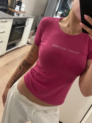 Rosa vintage Tommy Hilfiger tröja - Snygg vintage rosa t-shirt från Tommy Hilfiger med korta ärmar och rund hals. Framtill står det 'Tommy Hilfiger' i små glittriga stenar. Modellen är figurnära. Materialet är mjuk bomull. Storlek xs