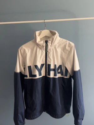 Helly Hansen jacka  - Snygg vindjacka i blått och vitt från helly hansen med stor logga över bröst och rygg. Jackan har dragkedja framtill, hög krage och elastiska muddar vid ärmsluten. Perfekt för blåsiga dagar och enkel att matcha till jeans.