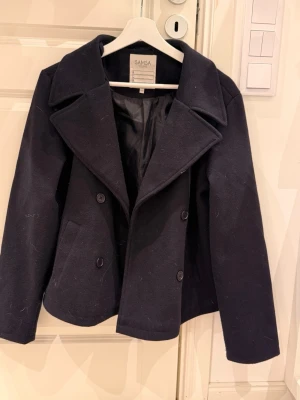 Marinblå kappa från SAMSA - Stilren marinblå kappa från SAMSA med breda slag, dubbelknäppning och två snedställda fickor framtill. Klassisk kort modell i ullblandning med helsvart foder. Perfekt för dig som gillar tidlös och enkel stil.