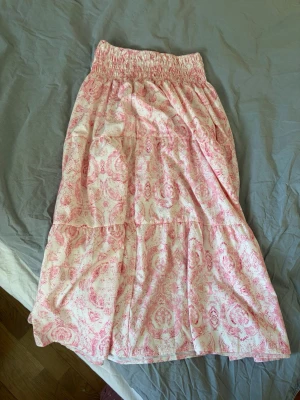 Långkjol med rosa mönster - Söt långkjol med vitt och rosa blommigt mönster och bred resår i midjan. Kjolen har en luftig och flowy passform, perfekt för varma dagar. Tillverkad i mjukt bomullstyg som känns skönt mot huden.