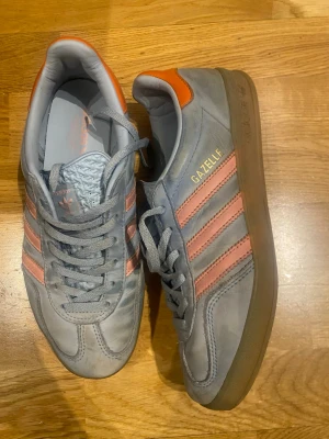 Adidas Gazelle ljusblå sneakers - Snygga Adidas Gazelle sneakers i ljusblå  mocka med klassiska rosa ränder och orange detalj vid hälen. Skorna har lågt skaft och gummisula. Perfekta för dig som vill ha en stilren och sportig look. 39 1/3 är storlek 