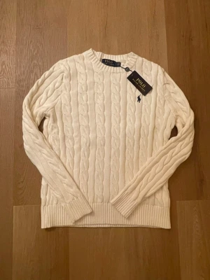 Vit kabelstickad tröja Polo Ralph Lauren - Kabelstickad tröja i mjuk vit ull från Polo Ralph Lauren. Klassisk rund halsringning, ribbade muddar och nederkant samt den ikoniska broderade loggan på bröstet. Perfekt för dig som gillar stilrena och tidlösa plagg.
