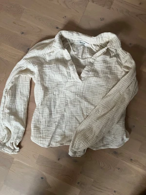 Krämvit blus från Kappahl i bomull - Luftig krämvit blus från Kappahl i mjuk, strukturerad bomullsmuslin. Blusen har v-ringning, avslappnad passform och långa ärmar. Perfekt för en chill och stilren look.