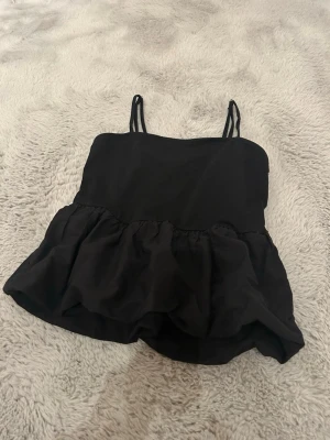 Svart volang topp - Snygg svart topp med smala axelband och volang nedtill. Toppen har en rak passform upptill, och en lite mer volangig/ puffugare passform nedtill, samt ärmlös design, perfekt för en trendig och enkel look. Materialet känns lätt och luftigt, vilket gör den skön att bära under varma dagar. Köpt för ca 1 år sedan, säljer du den ej kommer till användning, men fin och enkel att stajla. :))